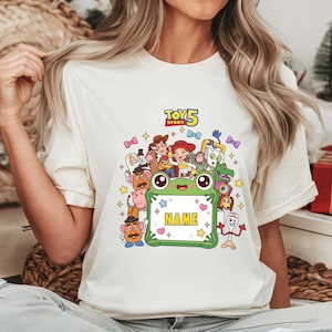May include: A cream-colored t-shirt featuring a colorful graphic of Toy Story characters surrounding a green frog-shaped frame with the text NAME in the center and the Toy Story 5 logo at the top, accented by small stars and bows.