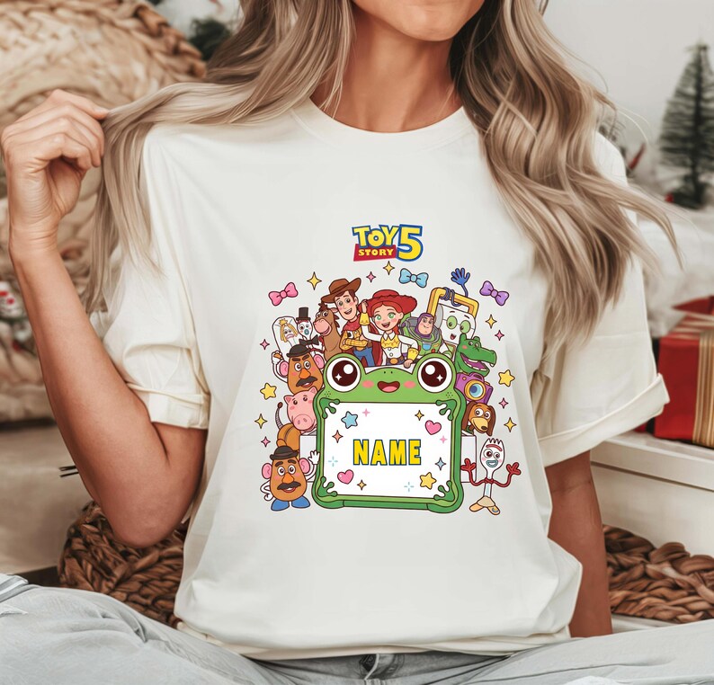 May include: A cream-colored t-shirt featuring a colorful graphic of Toy Story characters surrounding a green frog-shaped frame with the text NAME in the center and the Toy Story 5 logo at the top, accented by small stars and bows.