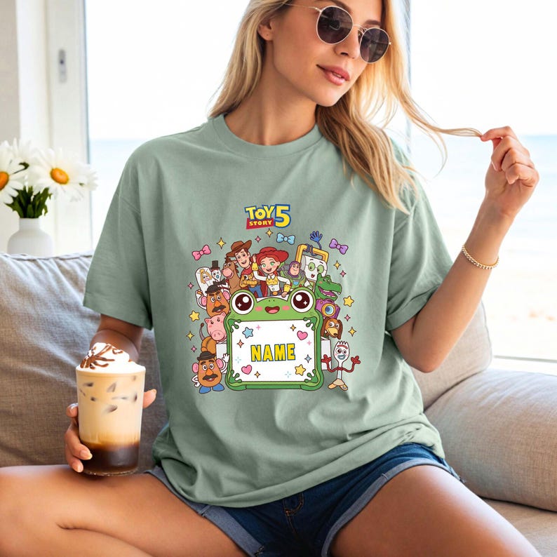 May include: A sage green t-shirt featuring a colorful graphic print of Toy Story 5 characters surrounding a central green frog-shaped frame with the word NAME printed in yellow. The design includes Woody, Jessie, Buzz Lightyear, and Forky.