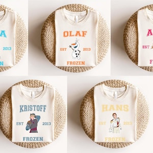 Puede incluir: Cinco camisetas blancas con diferentes personajes de Frozen impresos en ellas. Los personajes son Elsa, Olaf, Anna, Kristoff y Hans. Cada camiseta tiene el texto "EST" y "FROZEN" impreso en ella.