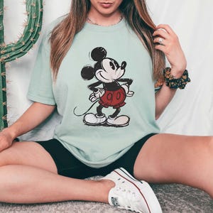 May include: A light green t-shirt featuring a classic Mickey Mouse illustration. The cartoon character is depicted in black, white, and red, with a smiling expression and hands on hips. The shirt is a relaxed fit.