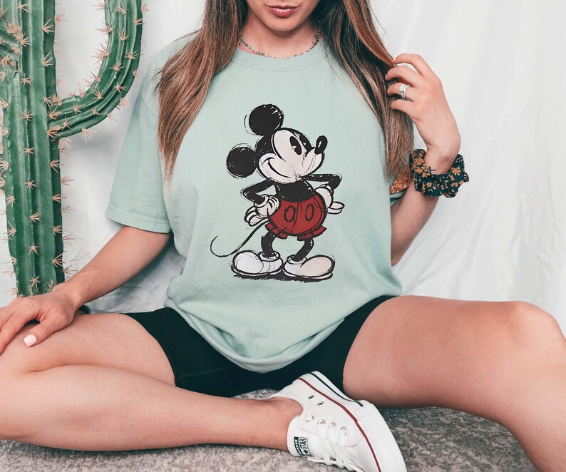 May include: A light green t-shirt featuring a classic Mickey Mouse illustration. The cartoon character is depicted in black, white, and red, with a smiling expression and hands on hips. The shirt is a relaxed fit.