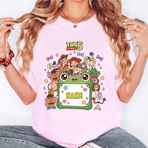 May include: A light pink t-shirt featuring a colorful graphic of Toy Story 5 characters surrounding a central green frog-shaped frame with the text NAME in yellow. The design includes Woody, Jessie, Buzz Lightyear, and other movie figures.