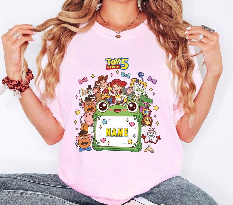 May include: A light pink t-shirt featuring a colorful graphic of Toy Story 5 characters surrounding a central green frog-shaped frame with the text NAME in yellow. The design includes Woody, Jessie, Buzz Lightyear, and other movie figures.
