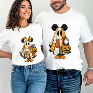 May include: White t-shirts featuring a Halloween-themed design. The design depicts a ghost character with Mickey Mouse ears, a patchwork-style outfit, and a jack-o'-lantern. The ghost is wearing orange shoes and holding a pumpkin.