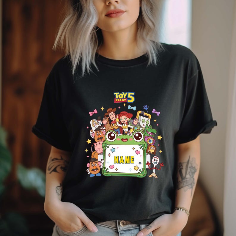 May include: A black short-sleeved t-shirt featuring a colorful graphic print of Toy Story 5 characters surrounding a green frog-shaped frame with the word NAME in the center. The design includes various animated figures and small star icons.