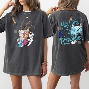 May include: Dark grey t-shirt with a colourful graphic of characters from the film Frozen, including Elsa, Anna, Olaf, and Sven. The back of the shirt has the text "Meet me in Arendelle" in blue script. A fun, casual tee.