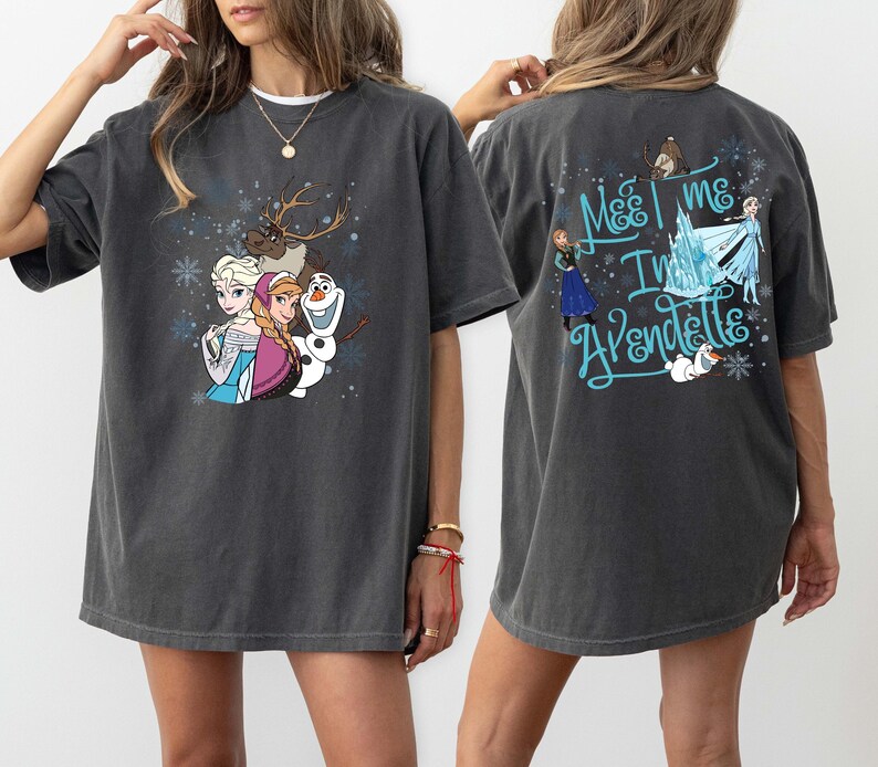May include: Dark grey t-shirt with a colourful graphic of characters from the film Frozen, including Elsa, Anna, Olaf, and Sven. The back of the shirt has the text "Meet me in Arendelle" in blue script. A fun, casual tee.
