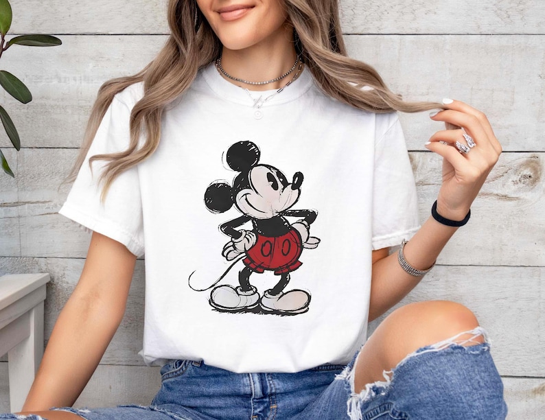 May include: White t-shirt featuring a classic Mickey Mouse graphic. The cartoon character is depicted in black, red, and white, with a vintage, sketched style. The shirt is a casual, short-sleeved design.