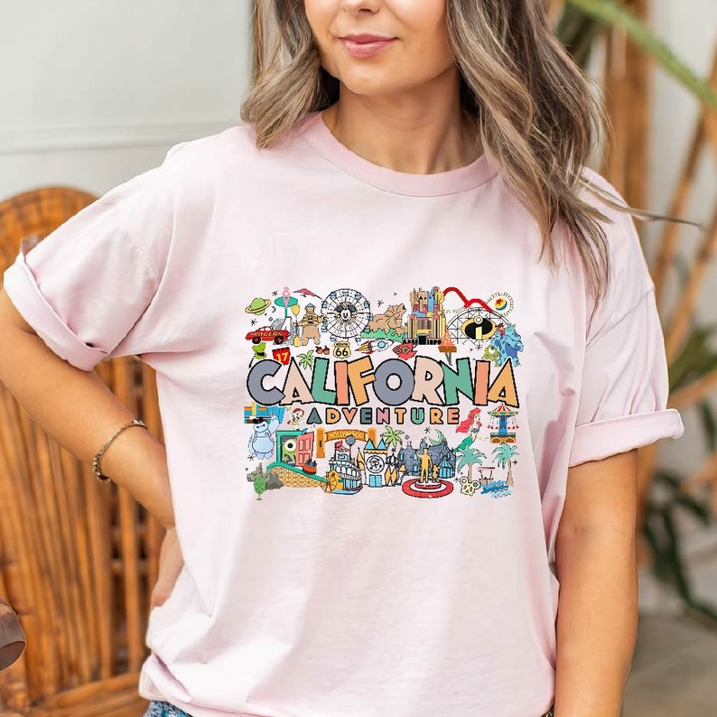 Pu&ograve; includere: Una maglietta rosa con un design grafico colorato che presenta il testo "California Adventure" e varie illustrazioni di monumenti e attrazioni della California.