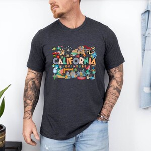 Pu&ograve; includere: Una maglietta grigio scuro con una stampa grafica colorata di monumenti californiani e il testo "California Adventure".