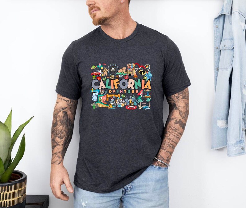 Pu&ograve; includere: Una maglietta grigio scuro con una stampa grafica colorata di monumenti californiani e il testo "California Adventure".