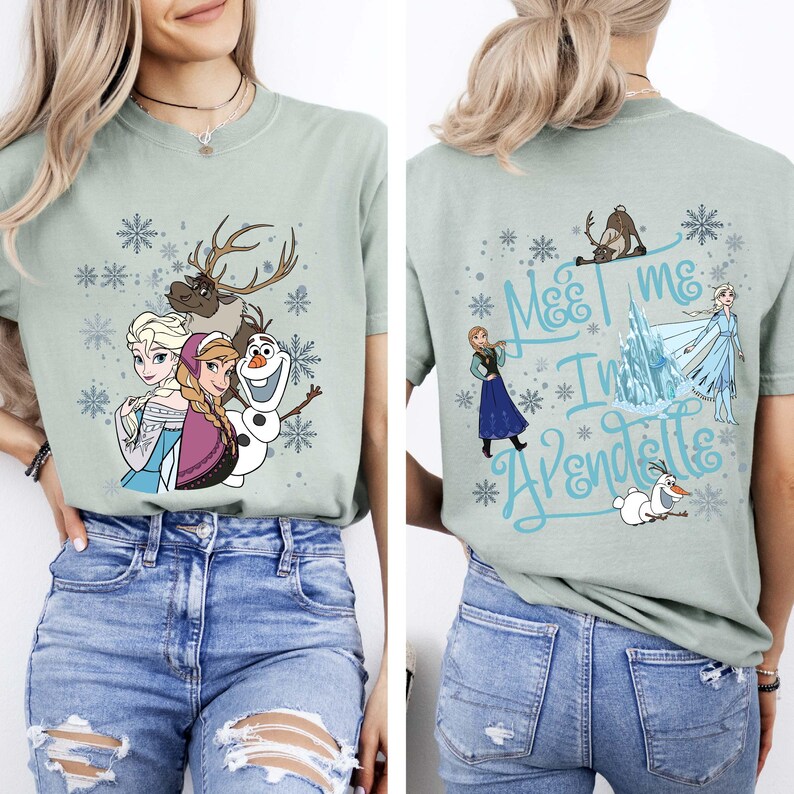 May include: Sage green t-shirt featuring characters from the movie Frozen, including Elsa, Anna, Olaf, and Sven, with snowflake accents. The back of the shirt has the text "Meet me in Arendelle" with additional characters.