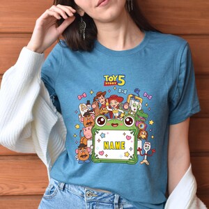 May include: A blue short-sleeved t-shirt featuring a graphic print of Toy Story 5 characters surrounding a green frog-shaped frame with the word NAME in yellow letters. The design includes Woody, Jessie, Buzz Lightyear, and other movie figures.