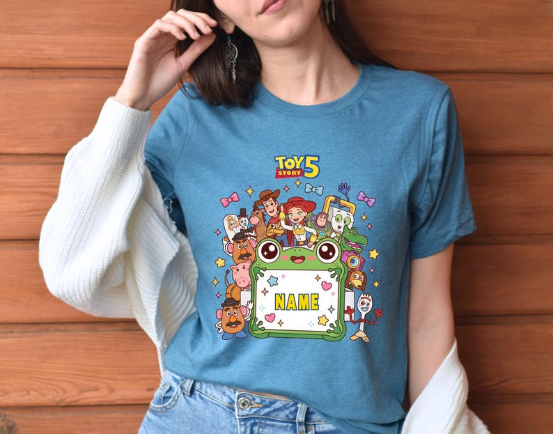 May include: A blue short-sleeved t-shirt featuring a graphic print of Toy Story 5 characters surrounding a green frog-shaped frame with the word NAME in yellow letters. The design includes Woody, Jessie, Buzz Lightyear, and other movie figures.
