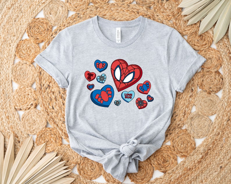 May include: A gray t-shirt with a graphic of a red and blue heart with a spider-man face on it. The heart is surrounded by smaller hearts with different designs. The text "Love Bug" is on one of the smaller hearts.