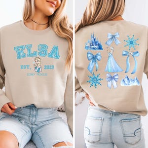 May include: Beige sweatshirt featuring the name "ELSA" in light blue block letters, with "EST. 2013" and a Disney Princess graphic. The back displays Elsa, bows, snowflakes, and a castle in shades of blue.