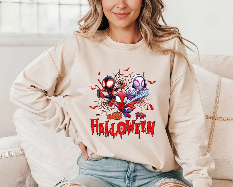 Peut inclure: Un sweat-shirt beige avec un imprim&eacute; graphique de trois personnages Spider-Man dans une toile. Les personnages portent des costumes rouge et bleu avec des masques noirs. Le texte "Happy Halloween" est imprim&eacute; en rouge sous les personnages.