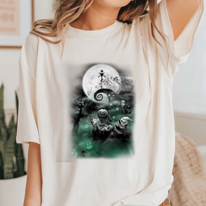 K&ouml;nnte beinhalten: Wei&szlig;es T-Shirt mit einem schwarz-wei&szlig;en Grafikdruck von Figuren aus dem Film Nightmare Before Christmas. Die Grafik zeigt Jack Skellington, Sally und Oogie Boogie vor einem Vollmond.