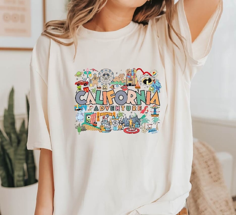 Pu&ograve; includere: Una maglietta bianca con un design grafico colorato che presenta il testo "California Adventure" e varie illustrazioni di monumenti e attrazioni della California.