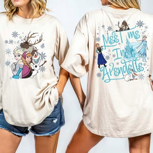 May include: Beige t-shirt featuring characters from the animated film Frozen. The front of the shirt displays Elsa, Anna, Olaf, and Sven. The back of the shirt has the text "Meet Me In Arendelle" with additional characters and snowflakes.