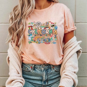 Pu&ograve; includere: Una maglietta rosa con un design grafico colorato che presenta il testo "CALIFORNIA ADVENTURE" e varie immagini di cartoni animati di attrazioni e punti di riferimento.