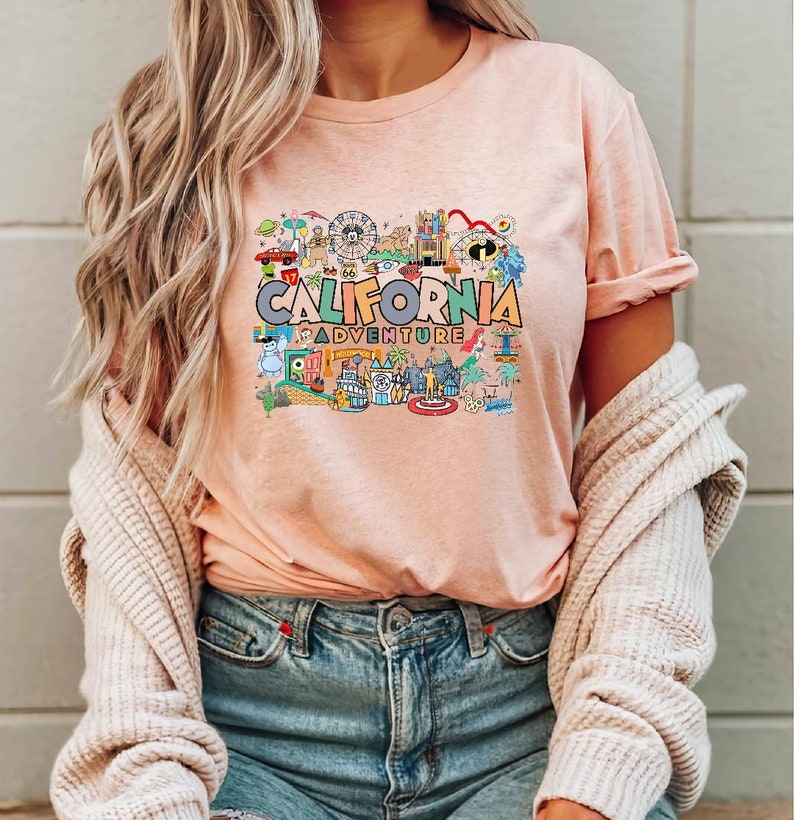 Pu&ograve; includere: Una maglietta rosa con un design grafico colorato che presenta il testo "CALIFORNIA ADVENTURE" e varie immagini di cartoni animati di attrazioni e punti di riferimento.