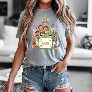 May include: A heather gray t-shirt featuring a colorful Toy Story 5 graphic. The design includes various characters surrounding a central green frog-shaped frame with the word NAME printed in yellow. Small stars and bows decorate the background.