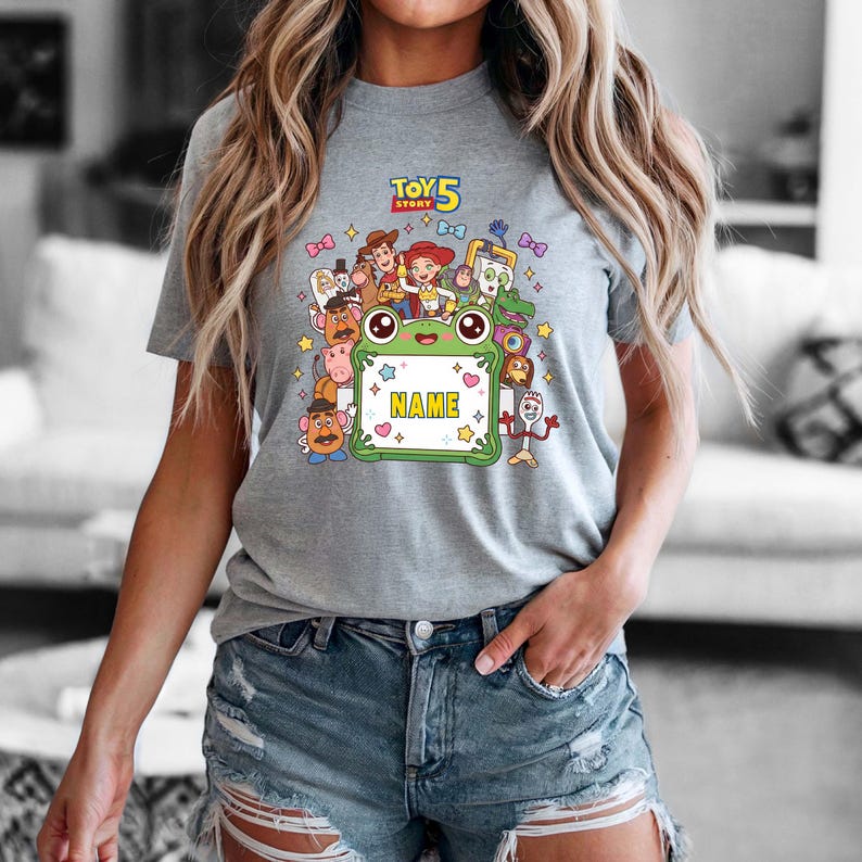 May include: A heather gray t-shirt featuring a colorful Toy Story 5 graphic. The design includes various characters surrounding a central green frog-shaped frame with the word NAME printed in yellow. Small stars and bows decorate the background.