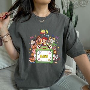 May include: A charcoal gray short-sleeve t-shirt featuring a colorful graphic print of Toy Story characters surrounding a central green frog-shaped frame with the text "TOY STORY 5" above and the word "NAME" inside the frame.