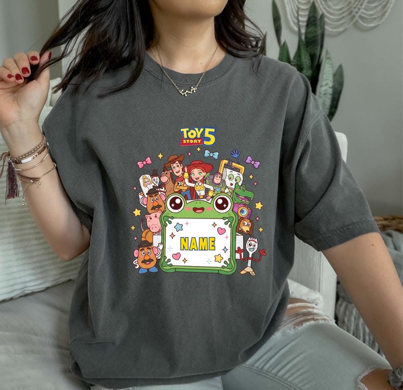 May include: A charcoal gray short-sleeve t-shirt featuring a colorful graphic print of Toy Story characters surrounding a central green frog-shaped frame with the text "TOY STORY 5" above and the word "NAME" inside the frame.