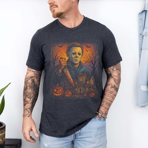 Może przedstawiać: Ciemnoszara koszulka z grafiką o tematyce Halloween. Projekt przedstawia postać z zakrwawionym nożem, dyniami, nawiedzonym domem i nagimi drzewami pod pomarańczowym niebem. Koszulka ma kr&oacute;tkie rękawy.