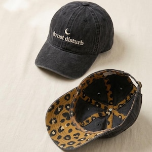 Puede incluir: Dos gorras de béisbol negras. Una gorra muestra el texto bordado "do not disturb" con una luna creciente. La otra gorra está al revés, revelando un forro con estampado de leopardo. Las gorras están hechas de un material suave y desgastado.
