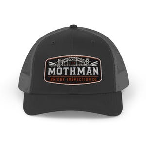 May include: A charcoal gray trucker hat with a mesh back and a solid front panel. An embroidered patch on the front features a bridge illustration, the text MOTHMAN, and the text BRIDGE INSPECTION CO. in white and orange thread on a black background.