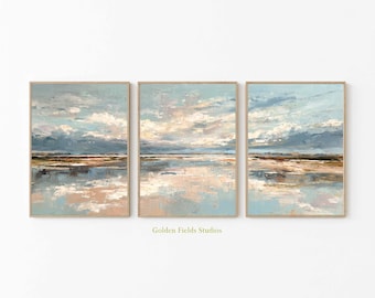 May include: Three framed landscape paintings with a serene coastal scene. The artwork features a palette of blues, beige, and peach, depicting a sky with clouds reflected in the water. The frames are a light wood color. The text "Golden Fields Studios" is at the bottom.