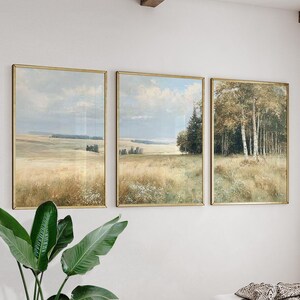 以下が含まれることがあります： 金色のフレームに入った3つの風景画。絵画は、畑、木、曇り空を描いています。アートワークは、自然の風景に焦点を当てた、落ち着いた色合いのヴィンテージな美学を持っています。絵画は白い壁に掛けられています。