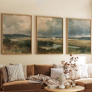May include: Three rectangular framed wall art prints featuring landscape paintings of a marshy field under a cloudy, overcast sky. The artwork displays muted tones of green, brown, and gray, depicting a natural horizon line with water.
