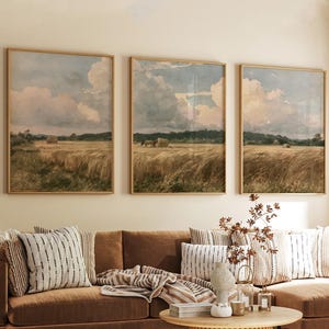 May include: A triptych of framed landscape prints featuring golden wheat fields under a cloudy sky. The three rectangular artworks are displayed side-by-side on a wall above a brown sofa, depicting rural scenes with hay bales and distant trees.