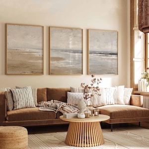 May include: A living room scene featuring a brown sofa with decorative pillows and a round wooden coffee table. Three framed beach landscape paintings hang above the sofa. A woven rug and a woven ottoman complete the cozy, neutral-toned space.