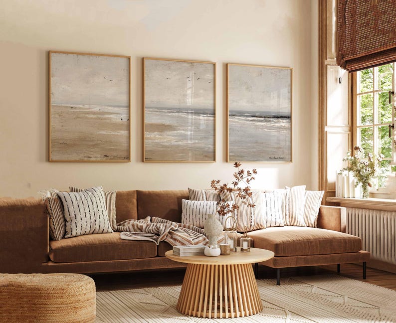 May include: A living room scene featuring a brown sofa with decorative pillows and a round wooden coffee table. Three framed beach landscape paintings hang above the sofa. A woven rug and a woven ottoman complete the cozy, neutral-toned space.