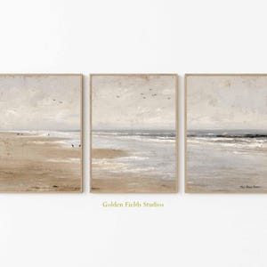 May include: Set of three framed beach landscape prints. Each print features a muted color palette of beige, gray, and blue, depicting a serene coastal scene with a sandy beach, ocean waves, and a cloudy sky. The frames are a light brown color.
