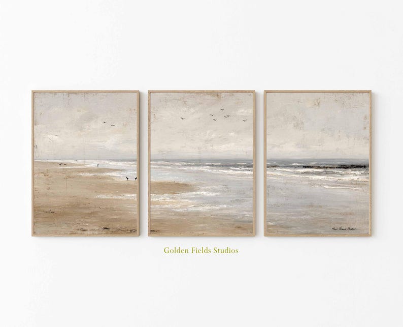 May include: Set of three framed beach landscape prints. Each print features a muted color palette of beige, gray, and blue, depicting a serene coastal scene with a sandy beach, ocean waves, and a cloudy sky. The frames are a light brown color.
