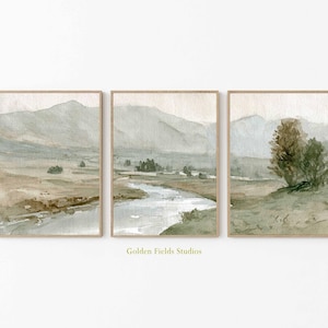 Könnte beinhalten: Set aus drei gerahmten Aquarell-Landschaftsdrucken. Jeder Druck zeigt eine ruhige Flussszene mit Bergen im Hintergrund. Die Farbpalette umfasst gedämpfte Grüntöne, Brauntöne und Blautöne. Der Text "Golden Fields Studios" befindet sich unten.