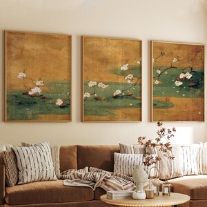 May include: Three framed art prints depicting a serene scene with white blossoms, green water, and a golden background. The artwork is displayed above a brown sofa with patterned pillows, creating a calming aesthetic.