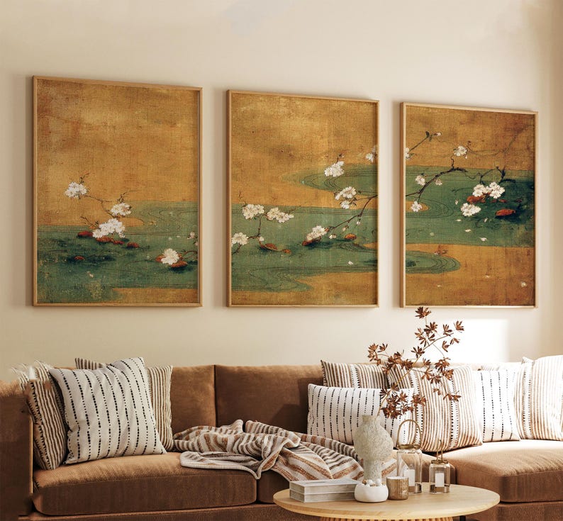 May include: Three framed art prints depicting a serene scene with white blossoms, green water, and a golden background. The artwork is displayed above a brown sofa with patterned pillows, creating a calming aesthetic.
