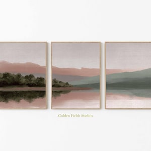 May include: Set of three framed landscape art prints. Each print features a serene landscape with a lake, trees, and mountains. The color palette includes muted tones of pink, green, and gray, creating a calming aesthetic. The text "Golden Fields Studios" is at the bottom.