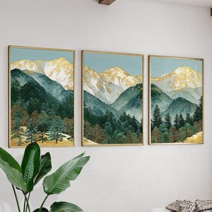 May include: A set of three framed landscape prints. Each print depicts a mountain range with gold-coloured peaks, green and teal-coloured forested slopes, and a light blue sky. The frames are gold-coloured.