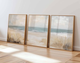 May include: Three framed abstract beach landscape paintings. Each artwork features a sandy beach, ocean, and sky in muted tones of beige, blue, and gray. The frames are a light brown color.