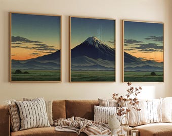 May include: Three framed landscape art prints depicting a mountain range at sunset. The artwork features a mountain with a snow-capped peak, green fields, and a colorful sky with shades of orange, blue, and yellow. The frames are a light brown color.