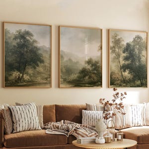 May include: Three rectangular framed wall art prints featuring landscape scenes of lush green trees, misty hills, and calm rivers. The artwork displays muted earth tones and soft lighting, presented in thin wooden frames hung on a plain wall.